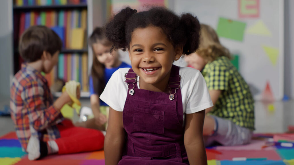 A young Black girl smiles while looking at the camera in a classroom.