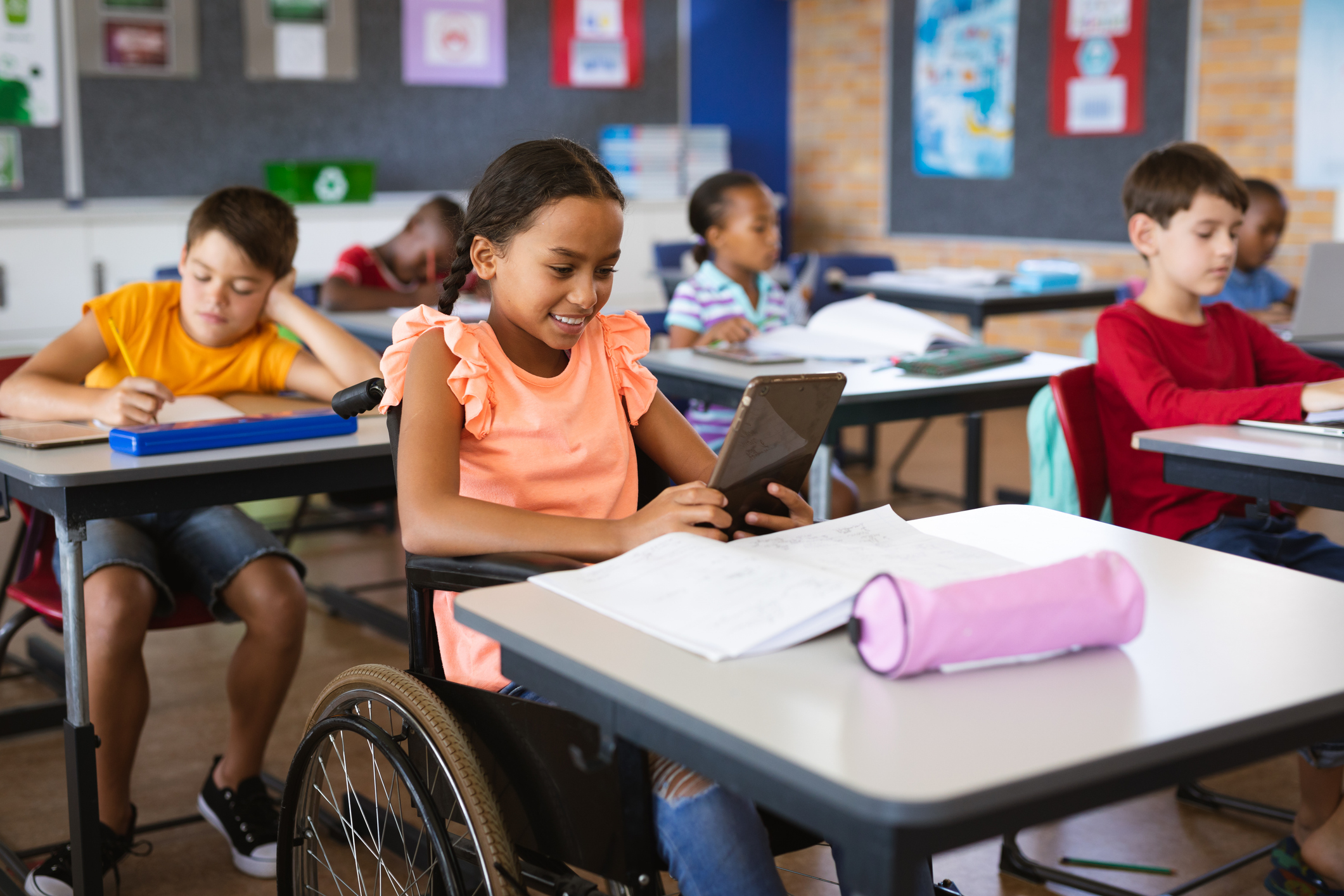 Decorative image of a female student looking at a tablet device in class. The student is in a wheelchair. Other students are also working at their desks.